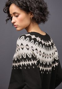 Sweter w czarno-białe wzory z geometrycznymi motywami, strukturalną tkaniną oraz okrągłym dekoltem. Prezentowany na modelce z kręconymi włosami.