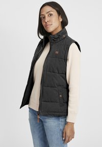 Svart quiltad väst med dragkedja, hög krage och tryckknappar. Bärs över en beigefärgad tröja med blå jeans. Logotypdetalj på bröstet.