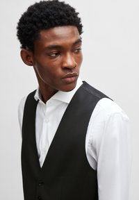 Svart vest over en hvit skjorte, med jevn tekstur, V-hals-design og frontknapper. Vestet har en satinaccent på skuldrene.