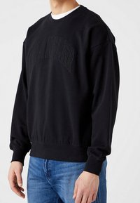 Felpa nera con colletto a girocollo e maniche lunghe, caratterizzata da un logo "Wrangler" in rilievo sulla parte anteriore. Tessuto morbido, vestibilità comoda.