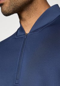 Σκούρο μπλε μπλουζάκι με μισό φερμουάρ και γιακά, με κομψή υφή, ελάχιστη ραφή και κοντό φερμουάρ. Το ύφασμα φαίνεται λείο και ελαστικό.