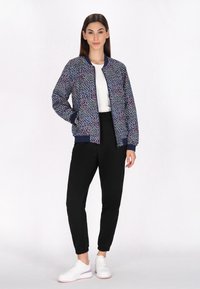 Jachetă bomber de culoare bleumarin cu un model floral colorat, cu manșete și tiv striate, purtată peste o cămașă albă și împerecheată cu pantaloni negri și adidași albi.
