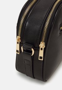 Czarna torba o teksturowanej powierzchni, wykonana z materiału syntetycznego, z podwójnymi złotymi zamkami błyskawicznymi, małym paskiem z akcentem w złotym kolorze oraz wyraźnie zarysowanym kształtem.