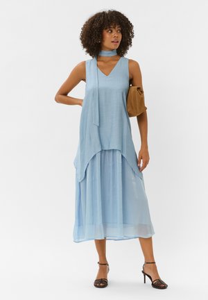 Femme aux cheveux bouclés portant une robe sans manches bleu clair avec une écharpe assortie, tenant une pochette marron et des sandales à talons noirs.