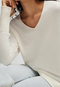 Frau in einem cremefarbenen V-Ausschnitt-Pullover und hellen, gewaschenen Jeans, stützt ihr Kinn entspannt auf ihre Hand.