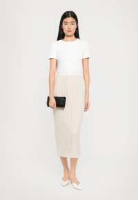Haut blanc à manches courtes associé à une jupe midi beige avec un motif texturé. Le mannequin tient une minaudière noire et porte des chaussures de couleur crème.