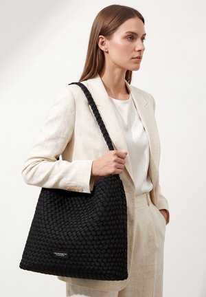 Femme en costume crème tenant un grand sac cabas noir tissé avec bandoulière, debout sur un fond neutre, regardant vers la droite.