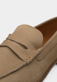 Mocassin en daim marron clair avec détails cousus et une lanière sur le dessus, doté d'un design sans lacets et d'une semelle en cuir lisse.