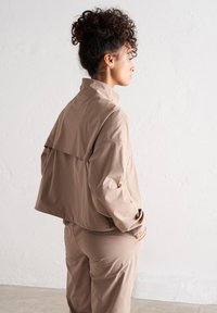 Beige krácená bunda s vysokým límcem a volným střihem, s dlouhými rukávy a vícevrstvým designem na zádech, doplněná o odpovídající kalhoty.