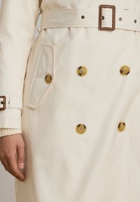 Light beige trench coat with a belt, featuring large round buttons and a side pocket. Smooth texture with a slight sheen.