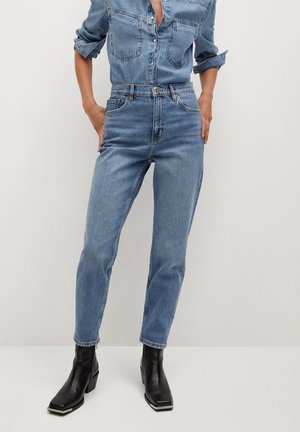 Persoon draagt lichtblauwe high-waist jeans, bijpassend spijkershirt met opgerolde mouwen en zwarte enkellaarsjes met hak, staand tegen een effen muur.