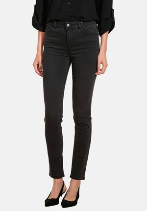 Donna che indossa jeans neri slim-fit con tasche borchiate e décolleté neri con tacco, vestita con una maglietta nera a maniche lunghe.