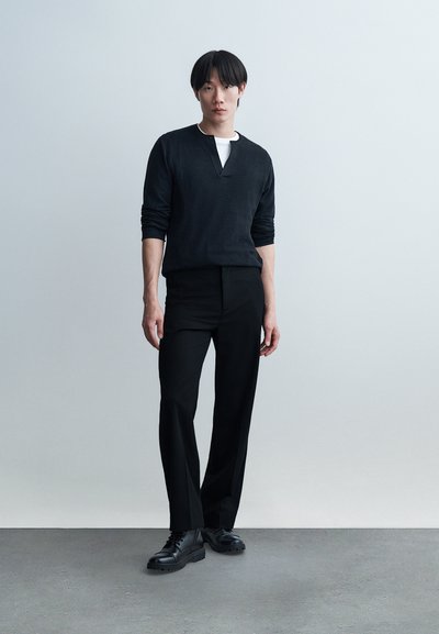 Νέος άνδρας όρθιος, φοράει μαύρο πουκάμισο με μακριά μανίκια πάνω από λευκό μπλουζάκι, μαύρο παντελόνι και μαύρα παπούτσια, μπροστά σε απλό γκρι ανοιχτό φόντο.