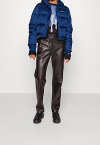 Niebieska kurtka puchowa z kapturem, czarny sweter z grafiką, brązowe spodnie z ekoskóry i masywne czarne buty z teksturowaną podeszwą.