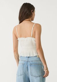 Bela smockirana kratka majica s volančki, tanka nastavljiva ramenska trakova, teksturirana tkanina, v kombinaciji z visokim modrim jeans kratkimi hlačami.