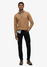 Pullover in maglia color nocciola con collo a mezza zip, indossato sopra una camicia a bottoni azzurro chiaro, abbinato a pantaloni neri e scarpe stringate marroni.