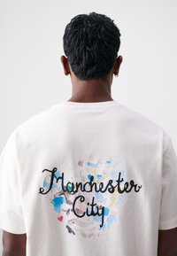 Krémové bavlněné tričko s barevným abstraktním grafickým potiskem a nápisem "Manchester City" v černém kurzívě na zadní straně. Krátké rukávy, standardní střih.