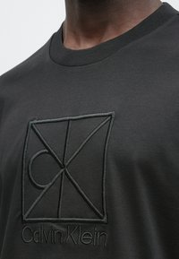 Μαύρο T-shirt με στρογγυλή λαιμόκοψη, με ανάγλυφο, κεντημένο λογότυπο σε μαύρο νήμα, κοντοκομμένο γύρω από την περιοχή του άνω στήθους.