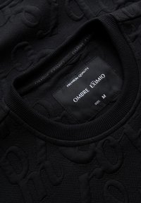 Pull à col rond noir texturé avec motif en relief et étiquette cousue indiquant "Premium Quality Ombre Eximio Taille M" à l'intérieur du col.