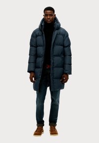Abrigo de plumón navy con un cuello alto, que presenta patrones de acolchado horizontales, llevado sobre un jersey negro de cuello alto, combinado con vaqueros azules y botas marrones.