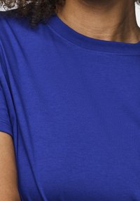Persona che indossa una maglietta a maniche corte di un blu brillante, con i capelli ricci parzialmente visibili nell'angolo superiore sinistro.