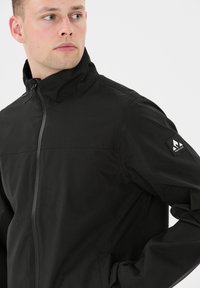 Svart jacka med hög krage, hel dragkedja och subtilt strukturerad tyg. Har en logotypapplikation på vänster ärm. Slimmad passform.