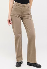 Beige Jeans mit hoher Taille, geradem Bein, dezenten Nähdetails und zwei Fronttaschen, kombiniert mit schwarzen Slippers.