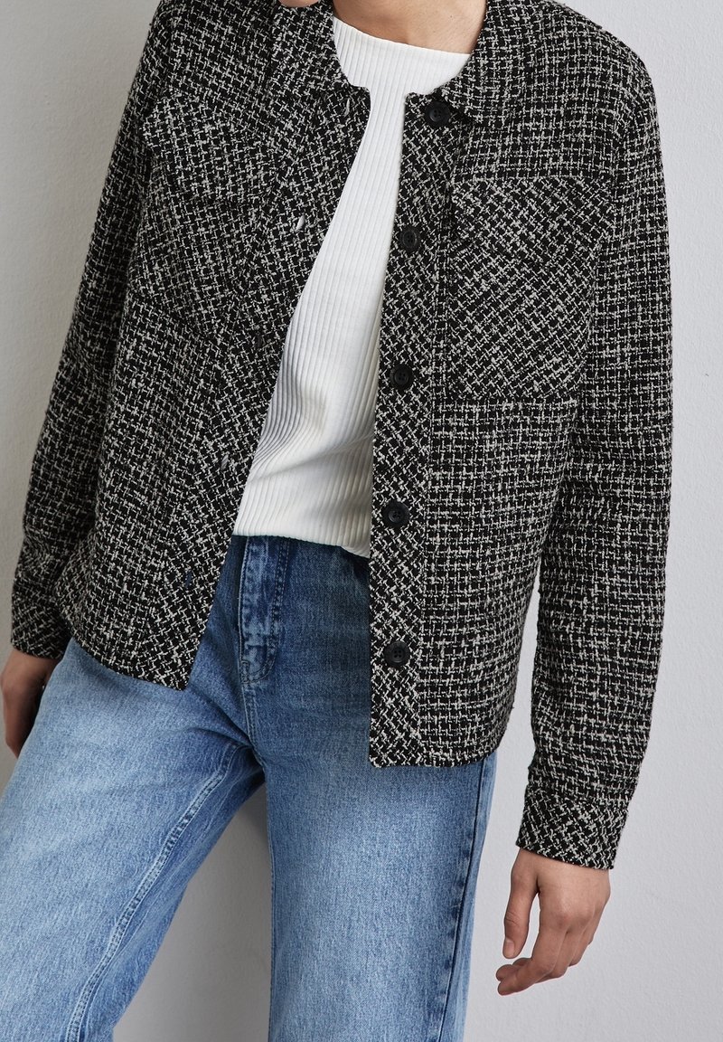 Zwart-witte geruite textuurjas met een kraag, knoopsluiting en twee voorkruiden, gedragen over een wit ribben shirt en lichtblauwe jeans.