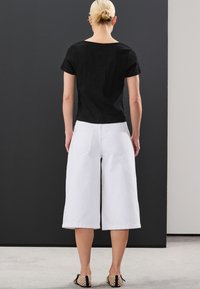 Mustast tekstuuriga lühikeste varrukatega topp, kombineeritud valgete laiade pükstega ja mustade libisevate jalanõudega, millel on lõhikuga detailid. Neutraalne taust.