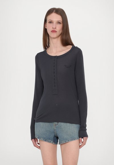 Camisa de manga larga azul marino con escote redondeado, cierre de botones en la parte delantera y logo bordado en el pecho. Combinada con pantalones cortos de mezclilla.