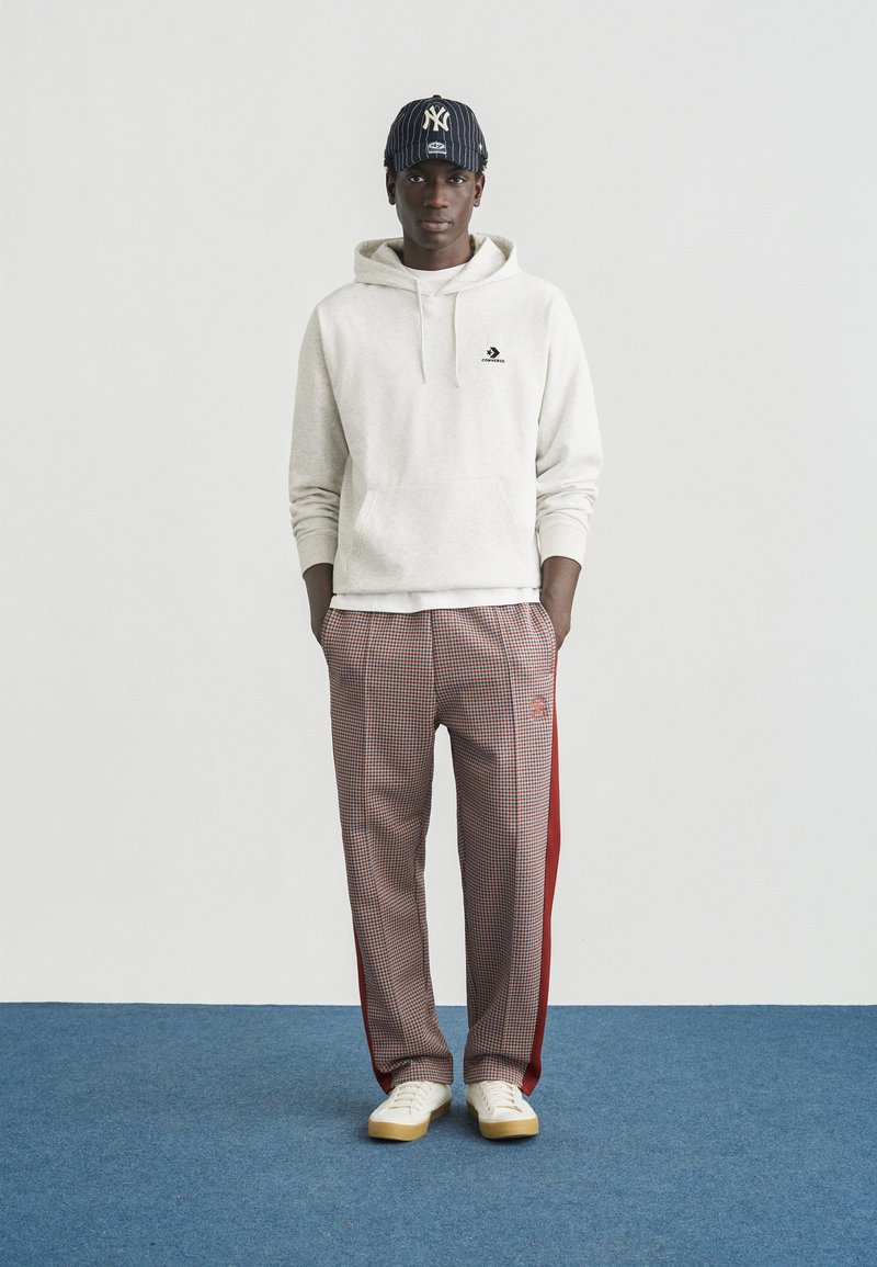 Sweat-shirt à capuche gris avec poche avant, pantalon à carreaux rouge et blanc avec bandes latérales, et baskets blanches sur un fond de tapis bleu.