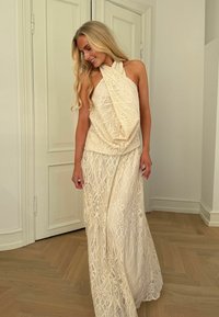 Femeie blondă, îmbrăcată într-un top bej din dantelă cu guler halter și o fustă lungă, stă în interior pe podea de lemn, privind în jos și zâmbind.