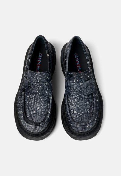 Black slip-on shoes with textured leather upper, featuring a raised pattern and thick rubber sole. Interior shows red and blue logo.