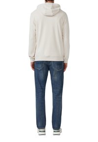 Hombre con sudadera blanca, jeans azules y zapatillas blancas, visto desde atrás sobre un fondo blanco.
