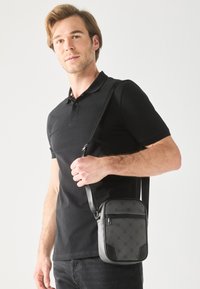 Black crossbody bag featuring a textured surface with a subtle pattern, adjustable strap, and a zippered front pocket.