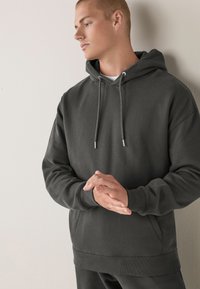 Dunkelgrauer Kapuzenpullover aus weicher Stoffqualität, versehen mit einer Kängurutasche und Zugbändern mit metallischen Enden; bequeme Passform.