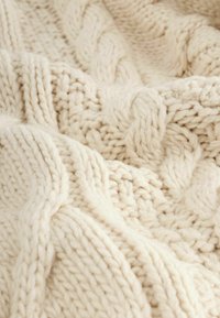 Cream knitted blanket with a cable pattern. Thick yarn creates a textured surface, featuring intertwining and smooth sections. Warm and cozy appearance.
