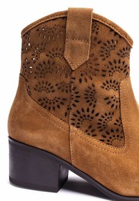 Brown suede ankle boot with cut-out floral pattern, square heel, and contrasting stitching along the seams. Textured finish, modern design.