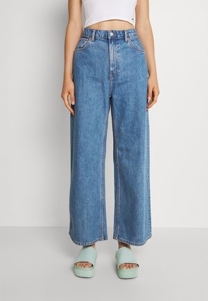 Person iført høj-taljede, vidde-ben blå jeans, hvid cropped tanktop og mintgrønne platform-slide sandaler, stående mod en ensfarvet baggrund.