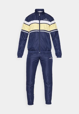 Marineblå treningsdrakt laget av glatt stoff har hvite og gule horisontale striper, høy krage, glidelås foran, og elastiske mansjetter og midjebånd.