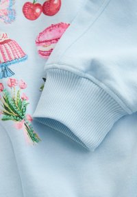 Sweat-shirt bleu clair avec des desserts et des fleurs brodés. Détail de poignets côtelés et texture douce avec des accents multicolores vibrants.