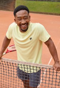 Lys gul idretts skjorte med korte ermer og en svart logo, sammen med grå shorts; lene seg mot et tennisnett med en racket.