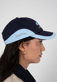 Femme aux longs cheveux foncés portant une boucle d'oreille en or et une casquette de baseball bleu marine et bleu clair, tournée vers la droite sur un fond gris.