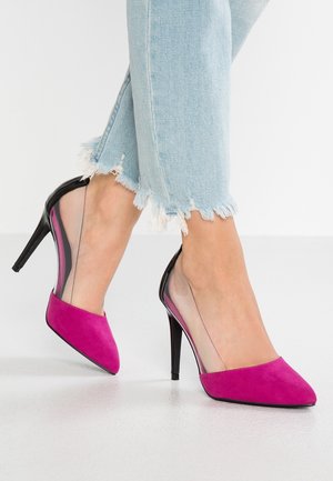 Žena nosící světle modré džíny s třepeněným lemem a růžové lodičky s průhlednými stranami na světlé podlaze.