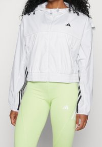 Femme portant une veste blanche zippée avec des bandes noires sur les manches et des leggings vert clair avec un logo blanc sur la cuisse.