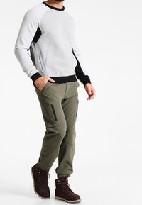 Grijze sweatshirt met zwarte accenten, gecombineerd met olijfgroene taps toelopende broek met ritszakken, en donkerbruine veterboots.