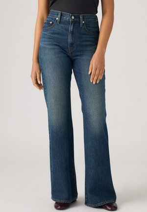 Personne portant un jean évasé bleu taille haute avec poches avant et fermeture à boutons, associé à un haut foncé et des chaussures bordeaux.