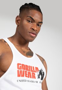 Bela majica na naramnice z napisom "GORILLA WEAR" v poudarjenem rdečem besedilu in grafiko gorile. Oblikovanje vključuje širok vratni izrez in gladko teksturo.