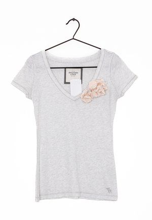 Jasnoszary, krótki rękaw, t-shirt z dekoltem w serek, z dwoma bladoróżowymi kwiatami z tkaniny przy dekolcie, zawieszony na czarnym wieszaku.