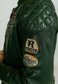 Veste en cuir vert avec un détail matelassé sur les épaules, ornée de deux patchs vintage : « RACING » et « REDSKINS SERVICE ». Poche zippée incluse.